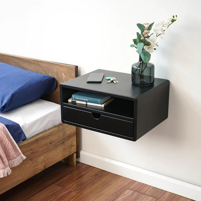 Black modern floating nightstand mounted on bedroom wall with books, keys, phone, and flower vase on top.