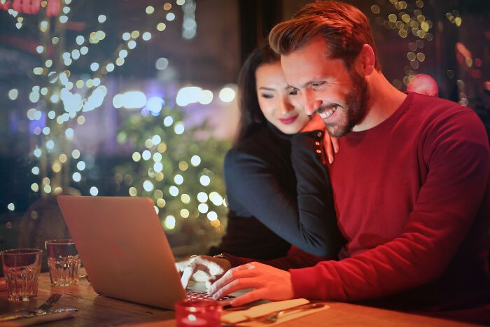 Couple smiling at laptop screen, possibly discussing shower thoughts in a cozy, festive environment.