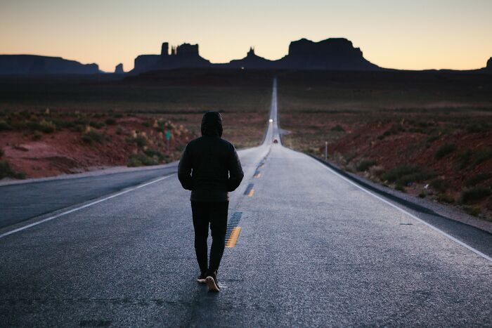 Person walking alone on a long road at dusk, possibly deep in shower thoughts, in a desert landscape setting.