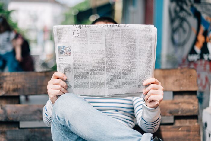 Person reading a newspaper on a wooden bench, pondering shower thoughts questions.