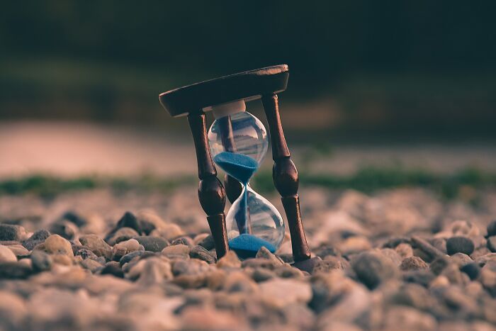An hourglass with blue sand on a bed of pebbles, symbolizing the passage of time and provoking shower thoughts questions.