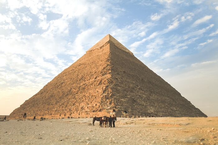A large pyramid with people and camels nearby under a clear blue sky, sparking shower thoughts questions about ancient structures.