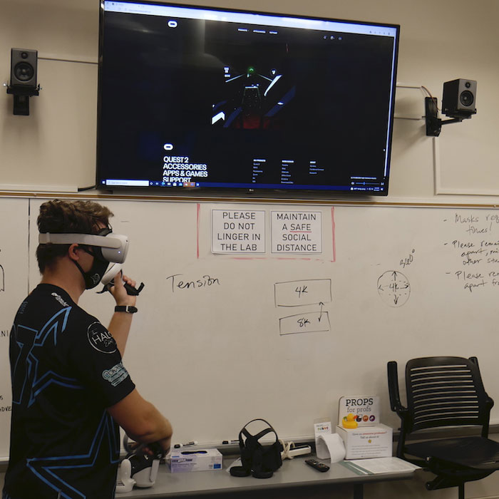 Person wearing a VR headset and mask in a game room with a scoreboard displayed on a large screen wall-mounted. Person wearing a VR headset and mask in a game room with a scoreboard displayed on a large screen wall-mounted.