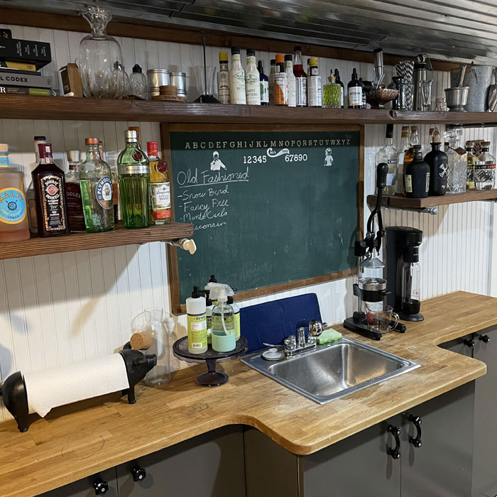 Small home bar shelving with assorted liquor bottles, glassware, and a chalkboard menu in a cozy wooden countertop space. Small home bar shelving with assorted liquor bottles, glassware, and a chalkboard menu in a cozy wooden countertop space.
