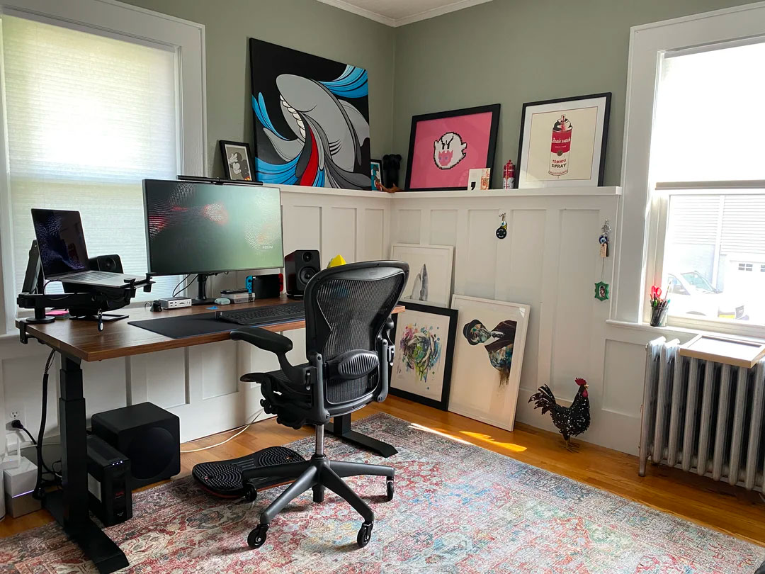 Home office with framed pictures displayed on a white wainscoted wall, featuring ergonomic chair and standing desk setup.