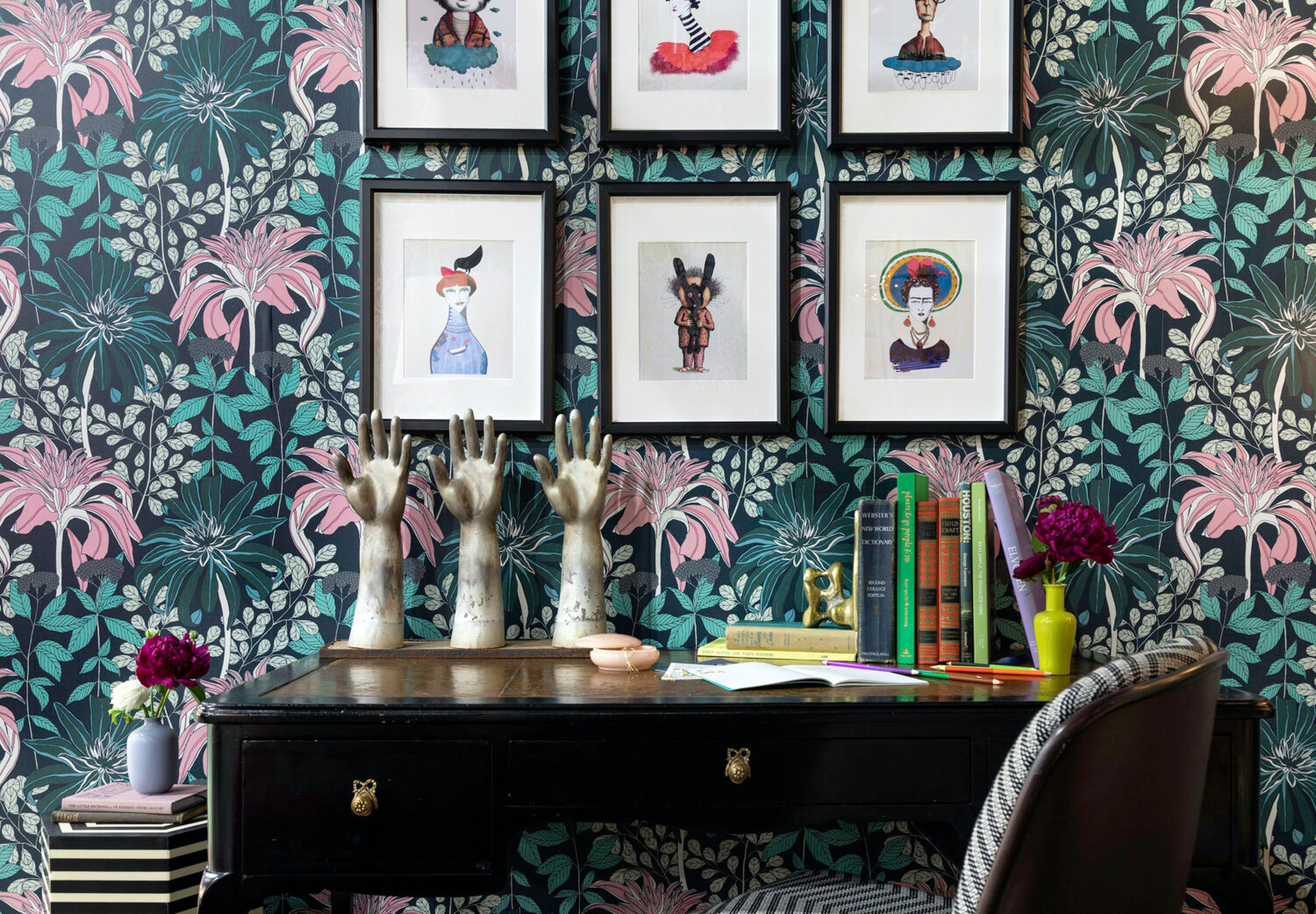Wooden table with artistic hand sculptures, colorful books, and framed whimsical art on a floral patterned wall for home office ideas.