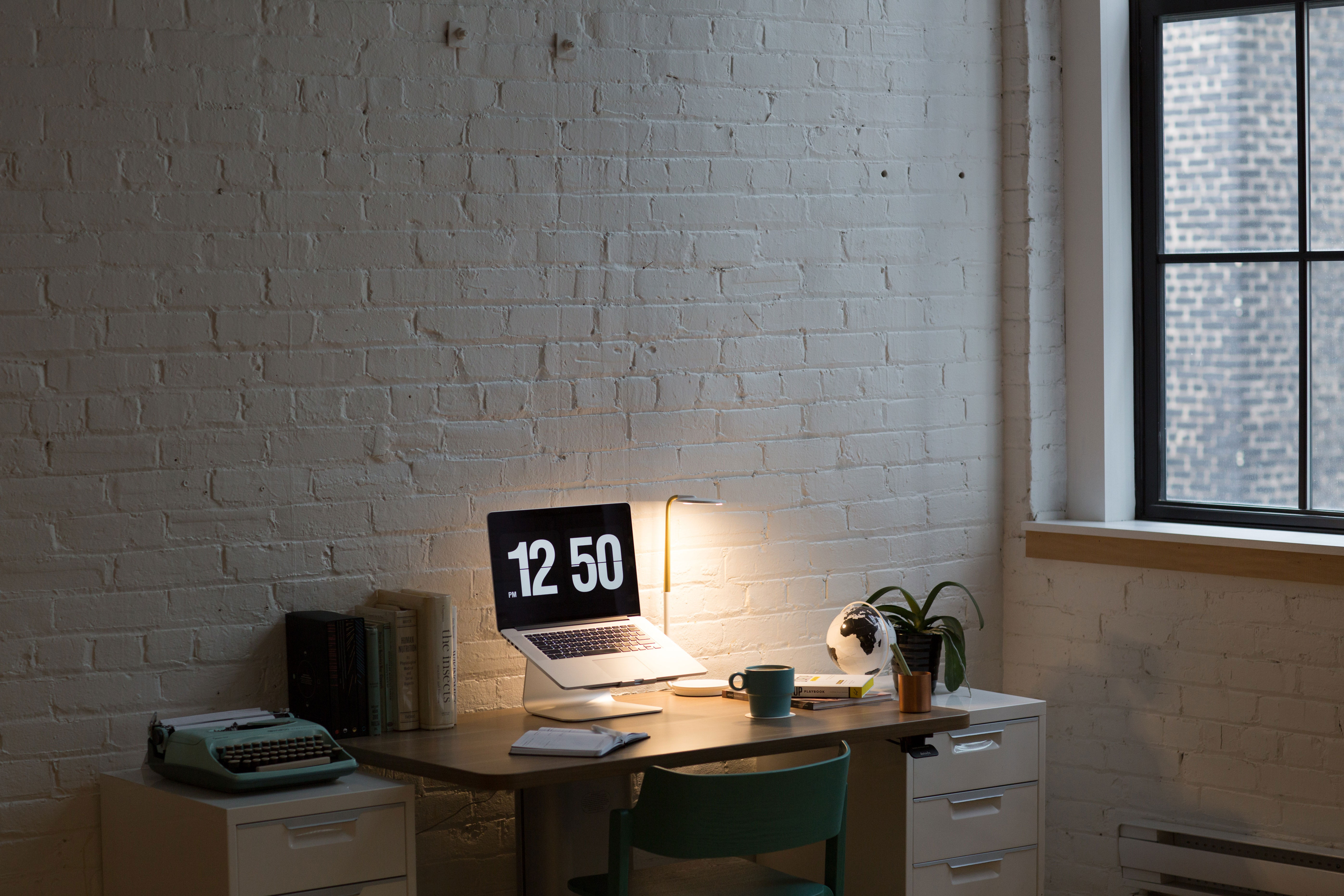 Minimal home office setup with MacBook turned on, desk lamp, books, and window light creating an inspired and productive workspace.