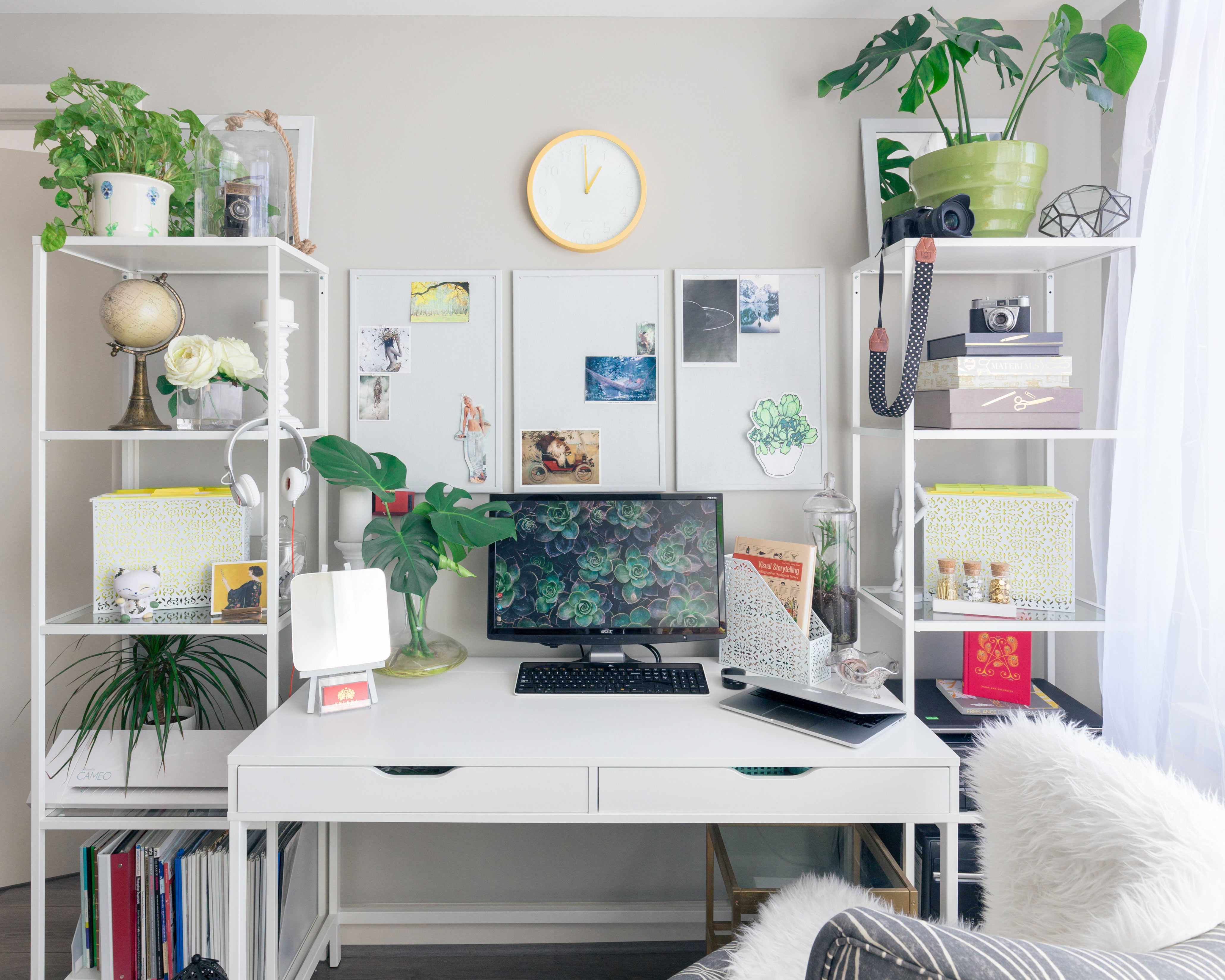 Bright home office setup with a turned on flat screen monitor on a white wooden desk surrounded by plants and shelves