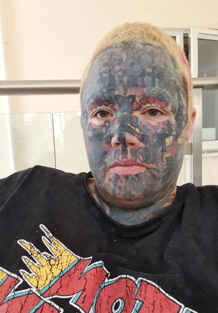 Woman with numerous tattoos, including facial tattoos, wearing a graphic tee, sitting indoors. Woman with numerous tattoos, including facial tattoos, wearing a graphic tee, sitting indoors.