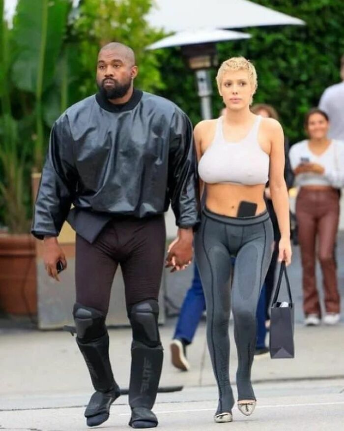 Kanye West with Bianca Censori wearing designer outfits, walking outside holding hands.