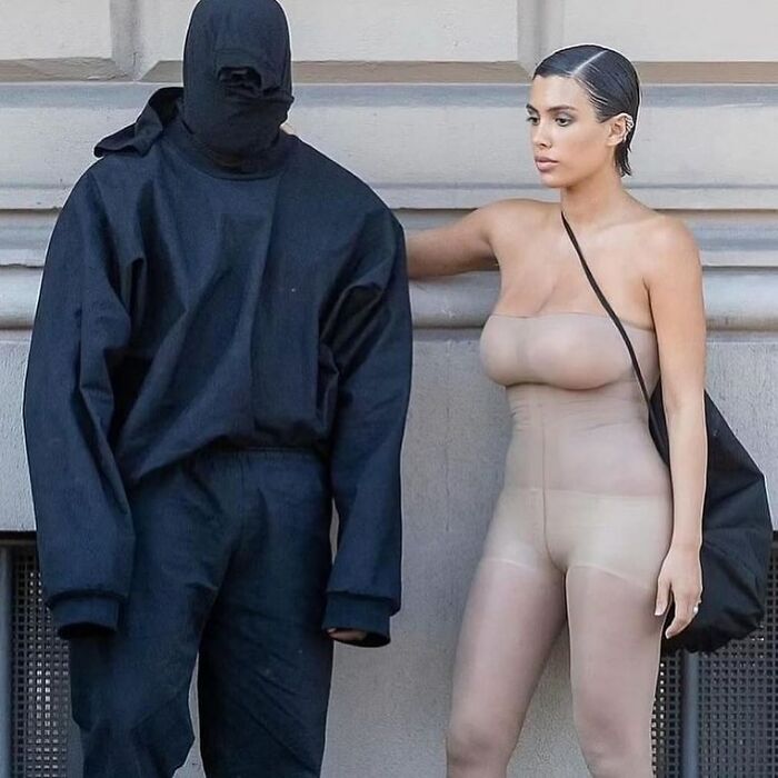 Two individuals standing, one in a black outfit with face covered, the other in a form-fitting outfit, against a concrete wall.