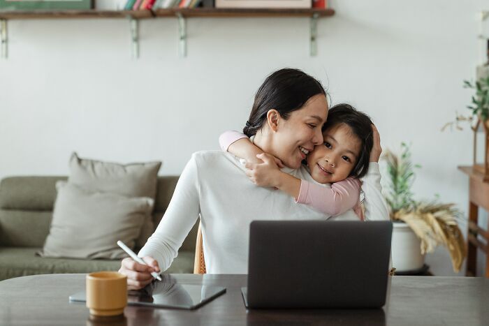 Mother and daughter sharing a warm hug while the mother works on a laptop at a cozy home workspace, highlighting unspoken mom code.