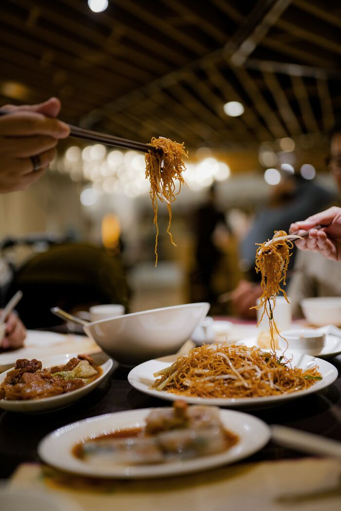 People using chopsticks to eat noodles and dishes at a restaurant, sharing food and culture in different countries.