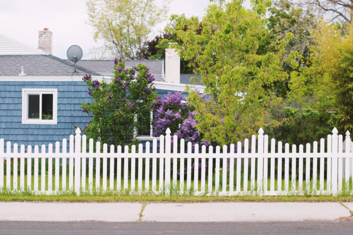 A white picket fence with a wave formation in front of a blue house and trees A white picket fence with a wave formation in front of a blue house and trees