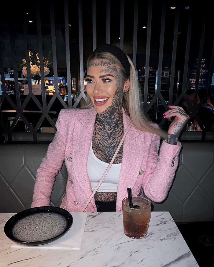 Influencer with fake forehead tattoo smiling in a pink jacket at a restaurant table.