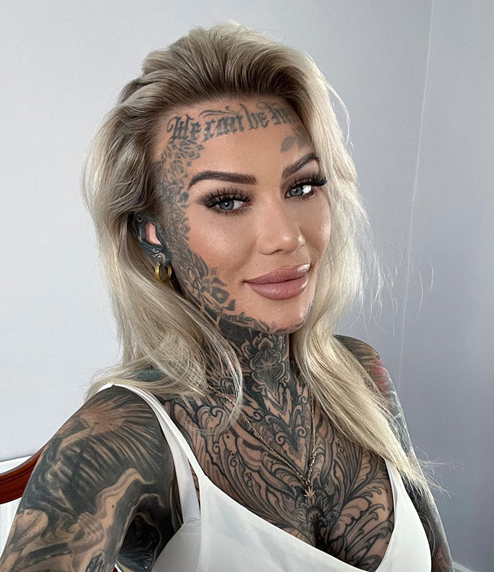 Influencer with tattooed forehead smiling, intricate facial and neck tattoos, long blonde hair, wearing a white top.