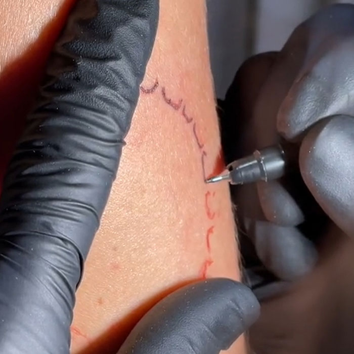 Tattoo artist inking arm, symbolizing love, using a tattoo machine while wearing black gloves.