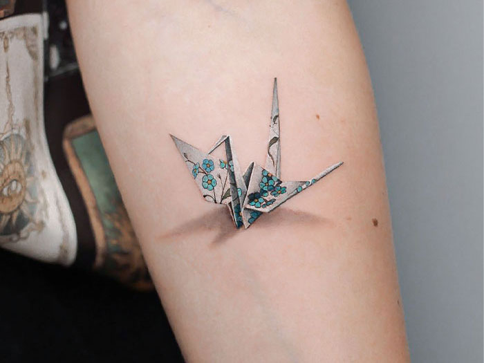 3D tattoo of an origami crane with floral patterns on an arm; realistic shadow adds depth.
