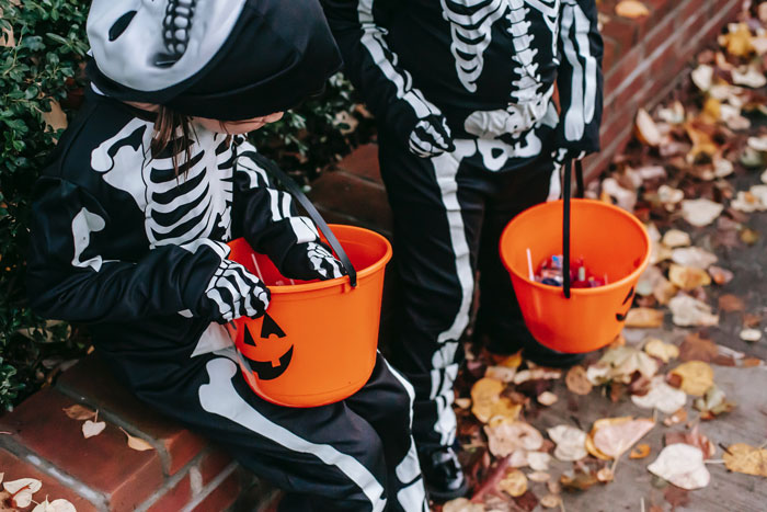Two children in skeleton costumes holding empty Halloween candy buckets outside on a leafy sidewalk. Two children in skeleton costumes holding empty Halloween candy buckets outside on a leafy sidewalk.