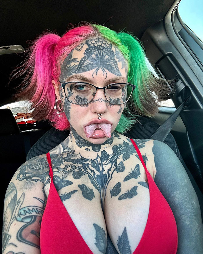 Tattooed woman with vibrant hair and facial piercings in a car, showcasing modified look.