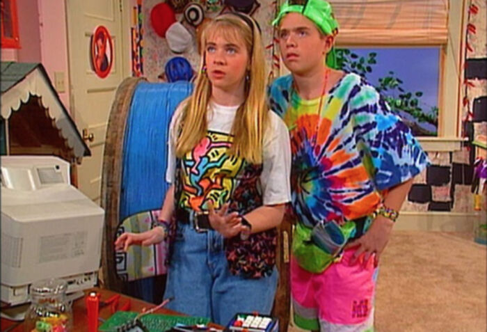Two kids in colorful '90s fashion looking surprised in a room with a computer.