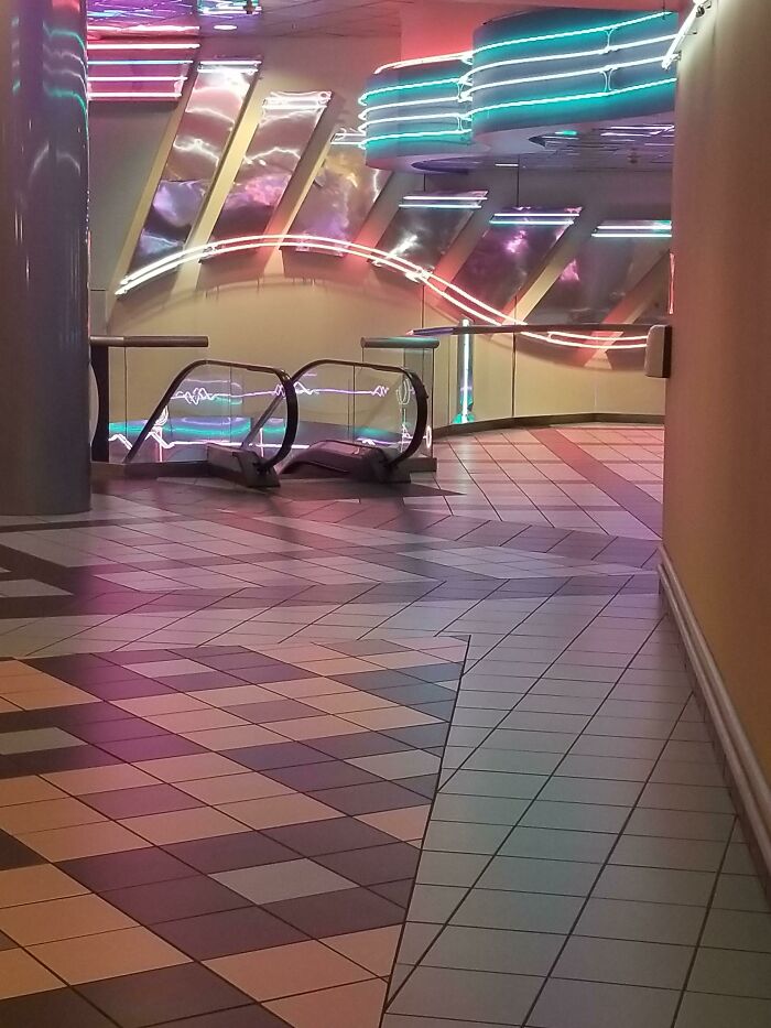 Neon-lit '90s mall interior with tiled floors and escalators, evoking nostalgia.