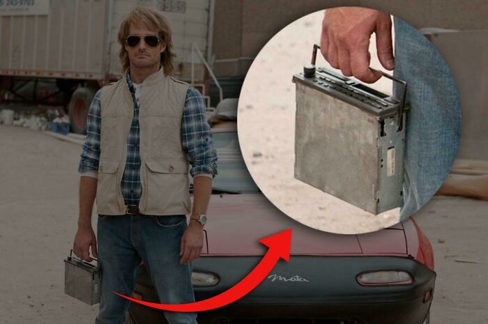 Man standing by a car with a car battery in hand, humorously illustrating feeling old faster than expected.
