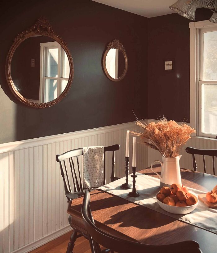 Dining room with dark walls, white wainscoting panels, wooden table, black chairs, and decorative mirrors reflecting natural light.