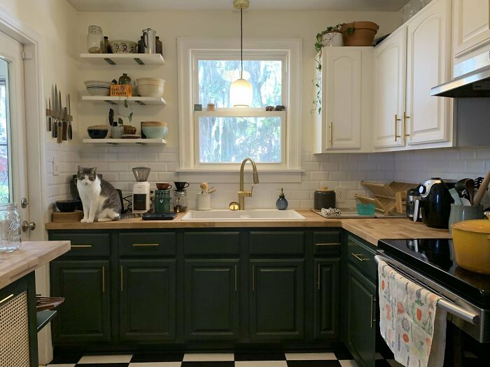 Kitchen with dark green kitchen cabinets and open wooden shelves, featuring a white farmhouse sink and gold faucet.