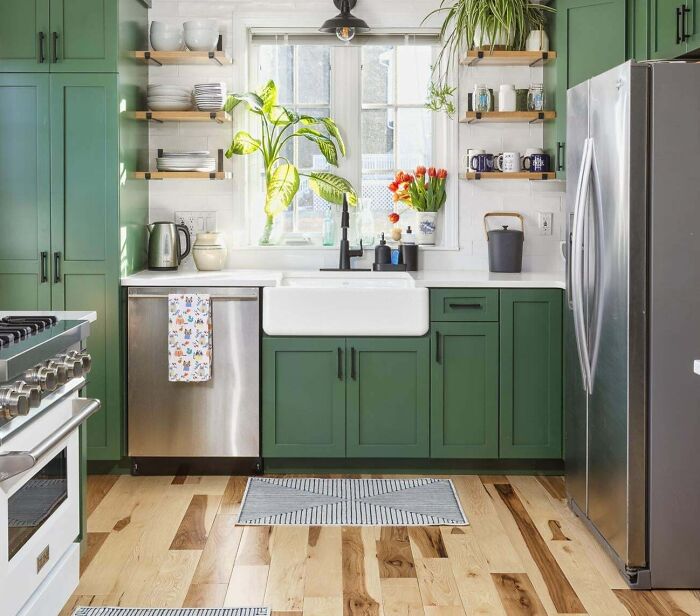 Green kitchen with green cabinets and open wooden shelves, featuring stainless steel appliances and natural light from a window.