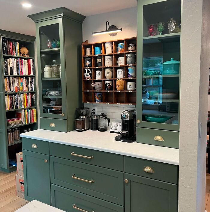 Green kitchen cabinets with glass doors and brass handles in a bright kitchen with organized coffee accessories.