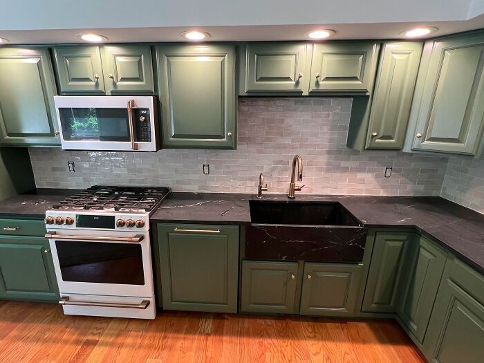 Green kitchen cabinets with brass fixtures, black countertop, and white appliances in a modern home kitchen design.