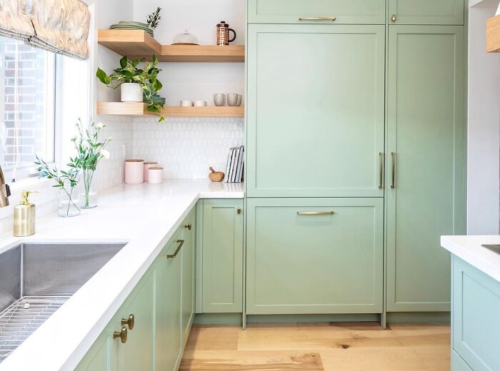 Light green kitchen cabinets with white countertops and wooden shelves in a bright modern kitchen space