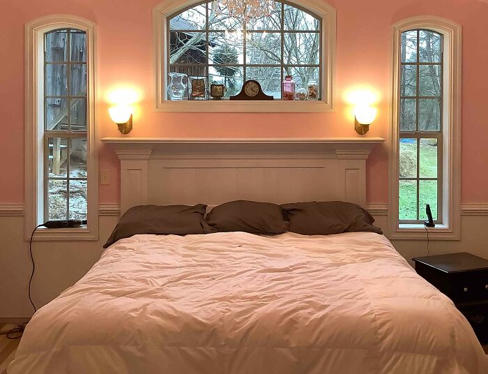 Pastel pink bedroom featuring a wainscoting panel headboard with symmetrical wall lights and large windows.