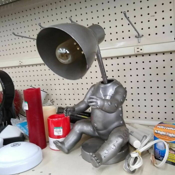 Weird thrift store lamp shaped like a baby doll with a lampshade head displayed among other donated items