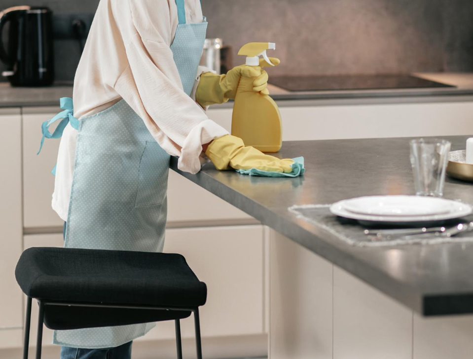 Woman wearing yellow gloves cleaning a concrete countertop in a modern kitchen with a spray bottle and cloth.