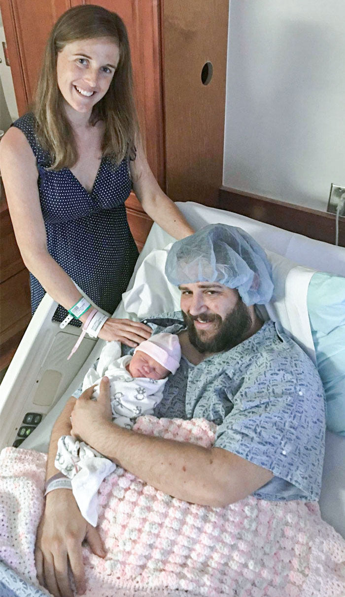Family photo in hospital: smiling man holding newborn, woman standing beside them, joyful moment.