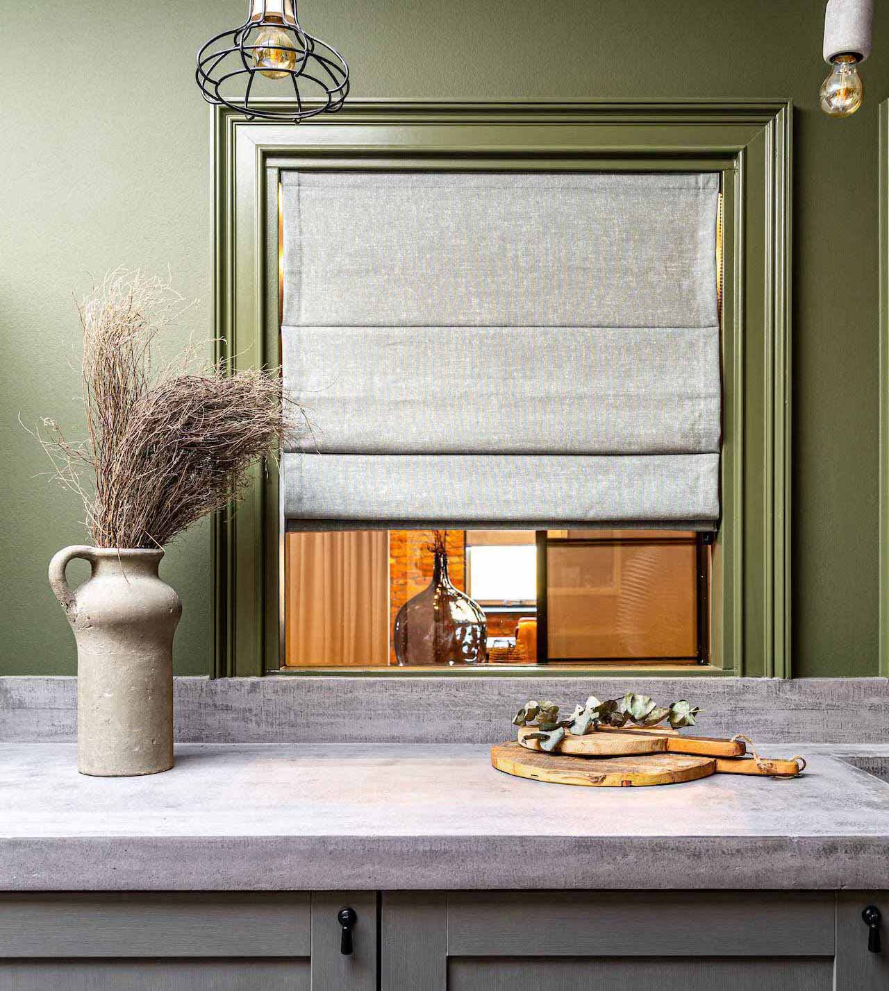 Rustic kitchen with rough concrete countertop, olive green walls, window with gray roman shade, and decorative vase with dried branches.