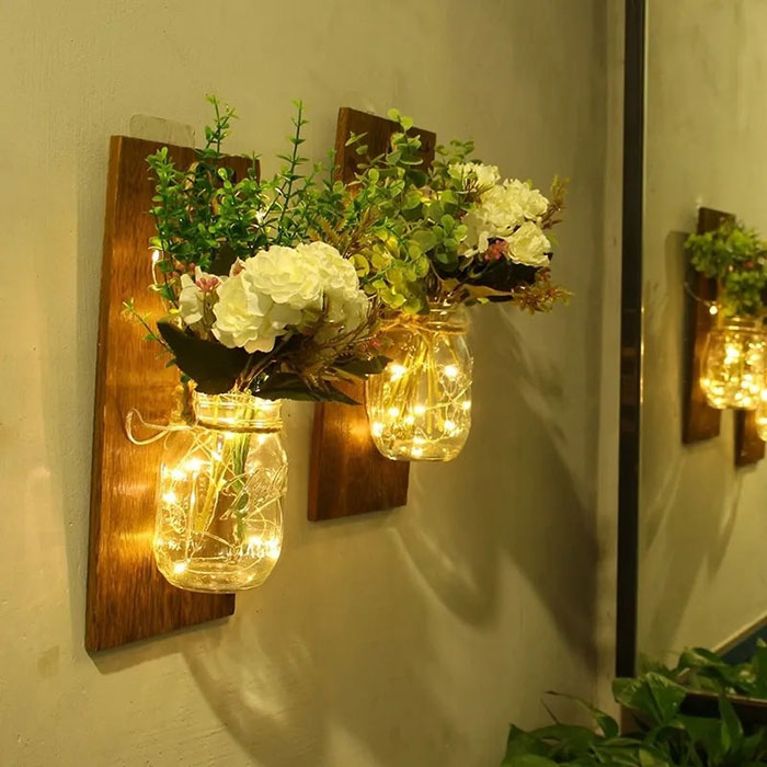 Two mason jars mounted on wooden wall plaques with fairy lights and flowers, a creative wall art idea for stylish spaces.