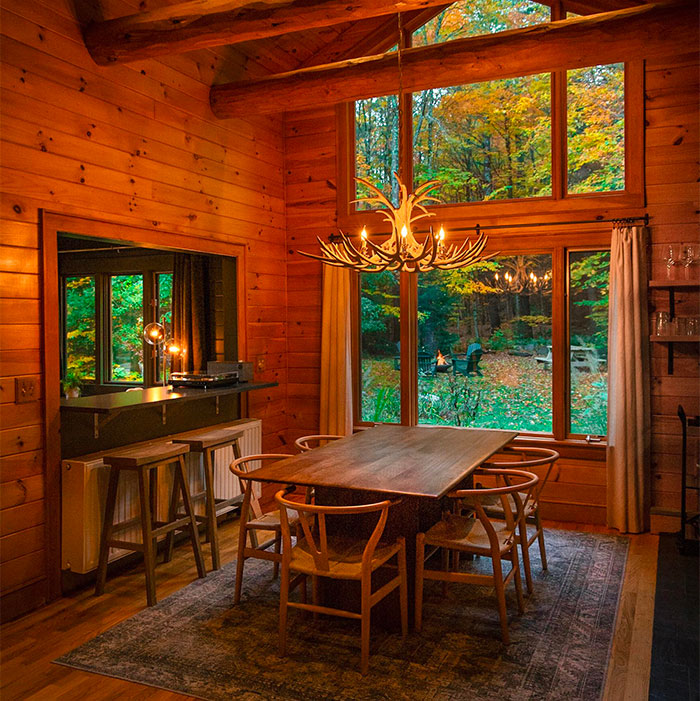 Cozy farmhouse dining room with wooden table and chairs, featuring large windows and warm autumn forest views.