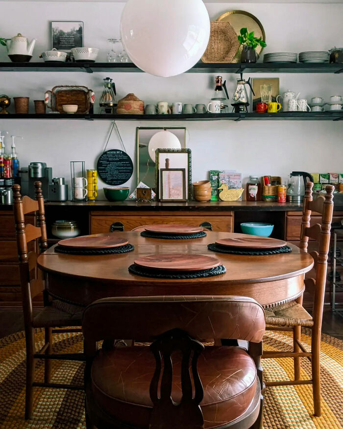Wooden dining table set with placemats in a cozy room featuring open shelves filled with kitchenware and decor.