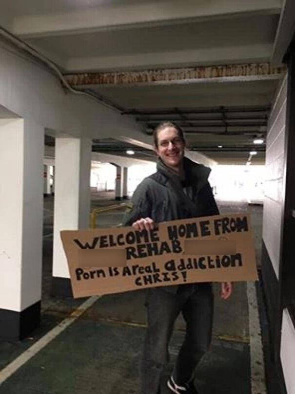 Man holding a funny airport pickup sign in a parking garage, smiling and holding a cardboard with humorous text.