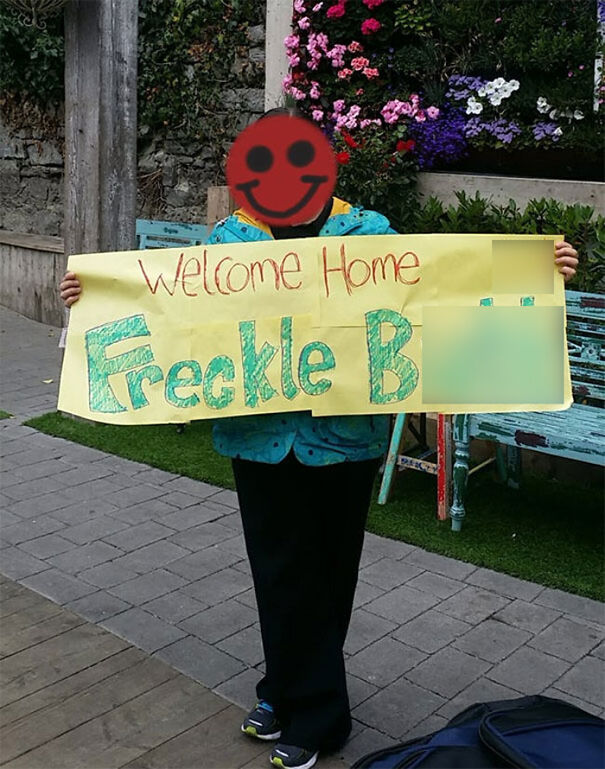Person holding a funny airport pickup sign with colorful text, standing near a flower wall.