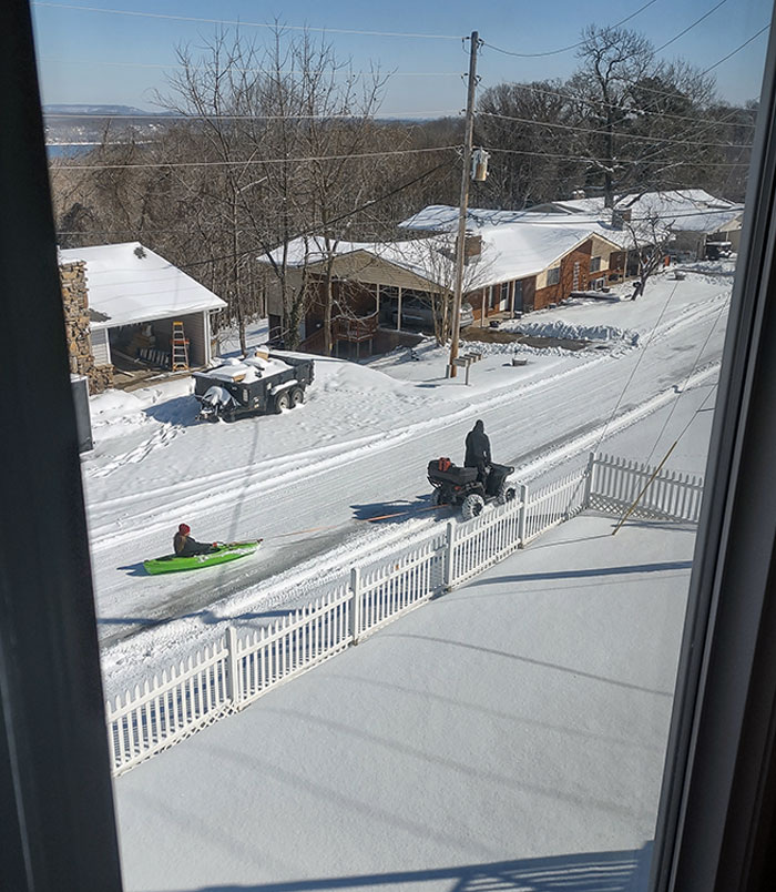 Parent with a sense of humor pulling a child in a kayak through snowy street using an ATV.