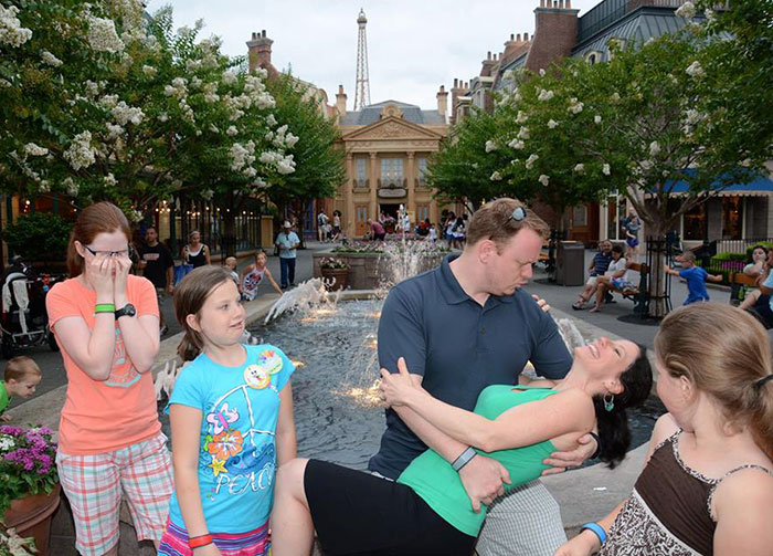 A parent playfully dips their partner near a fountain, while kids react amusedly in a park setting, showcasing parental humor.