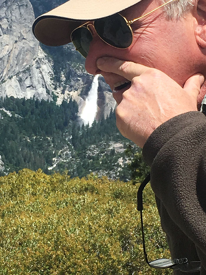 Man humorously posing with waterfall in background, wearing sunglasses and a cap, showcasing a playful parent's sense of humor.