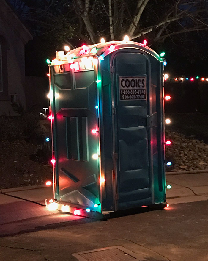 Portable toilet humorously decorated with colorful Christmas lights in a yard.