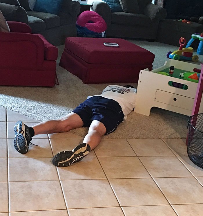 A parent humorously lying on the floor, head under a child's play table, showcasing their fun sense of humor.