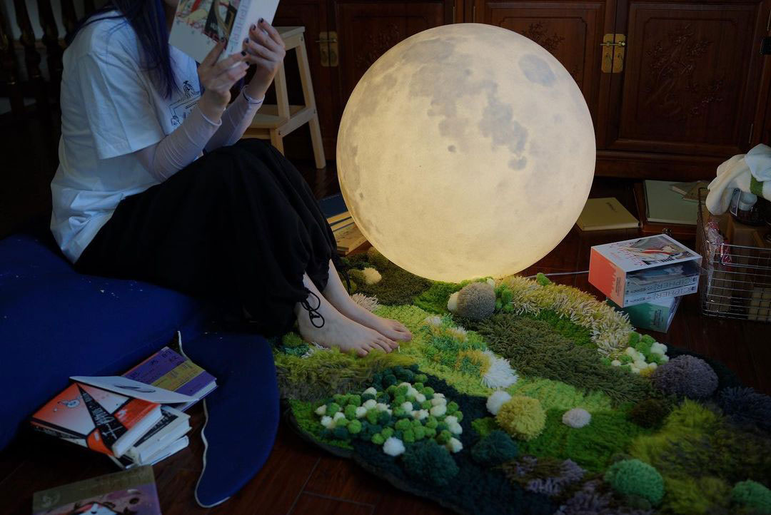 Woman sitting with feet on a textured moss rug next to a large glowing moon light in a cozy indoor setting Woman sitting with feet on a textured moss rug next to a large glowing moon light in a cozy indoor setting