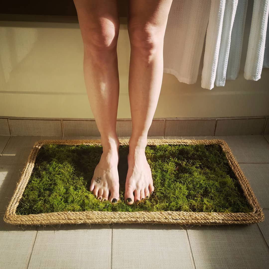 Woman standing barefoot on a natural moss bath mat framed with braided rope in a sunlit bathroom setting. Woman standing barefoot on a natural moss bath mat framed with braided rope in a sunlit bathroom setting.